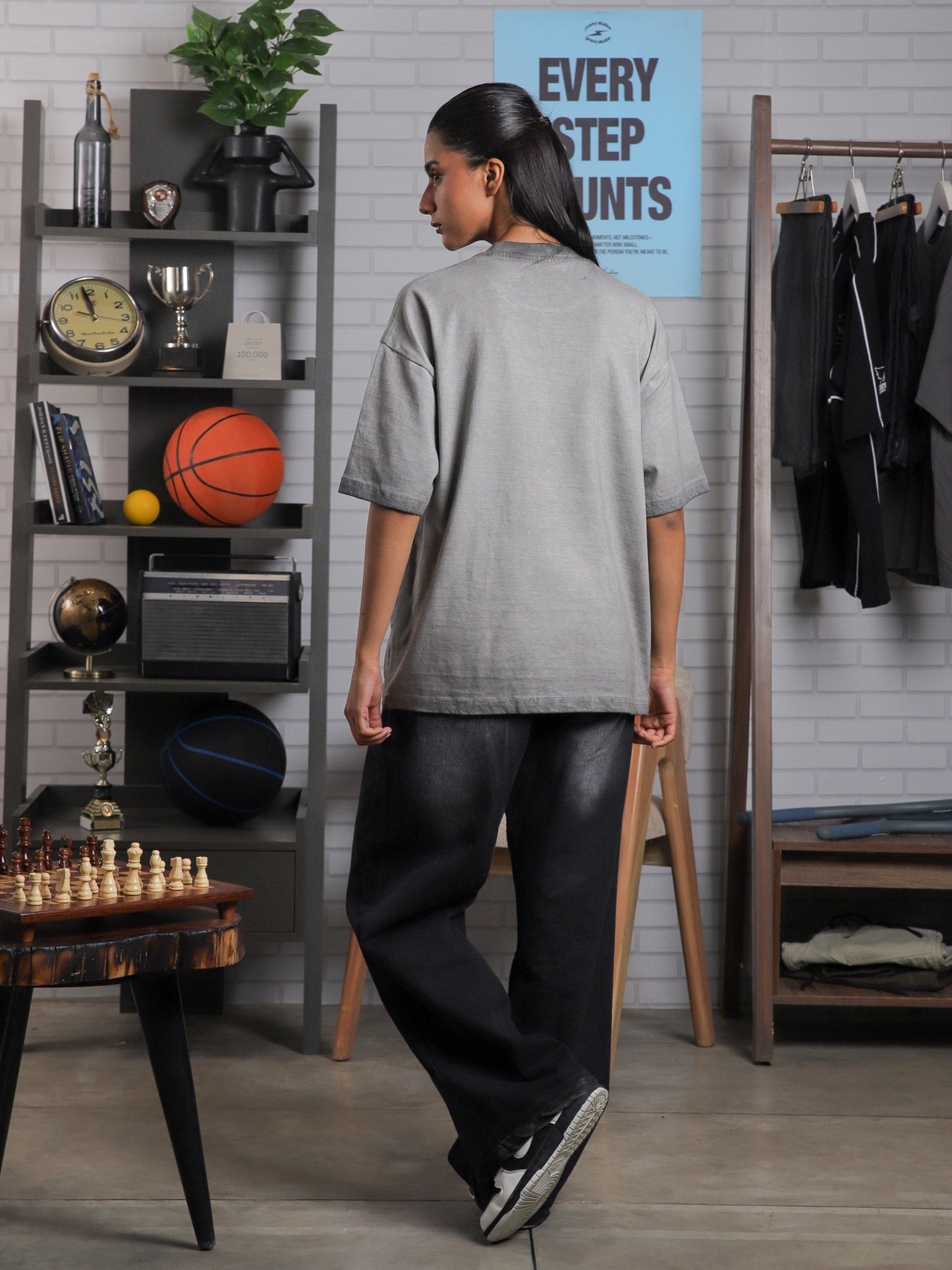 Female model turning away in Cultured Visionaries tee, set in a creative studio with decor including a shelf, basketball, and posters.