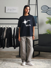 Front view of a woman wearing the navy "Never Stopping Culture" t-shirt with bold white star and CLTR logo, styled with grey cargo pants and white sneakers, standing in a minimal studio setup with clothing racks and indoor plants.
