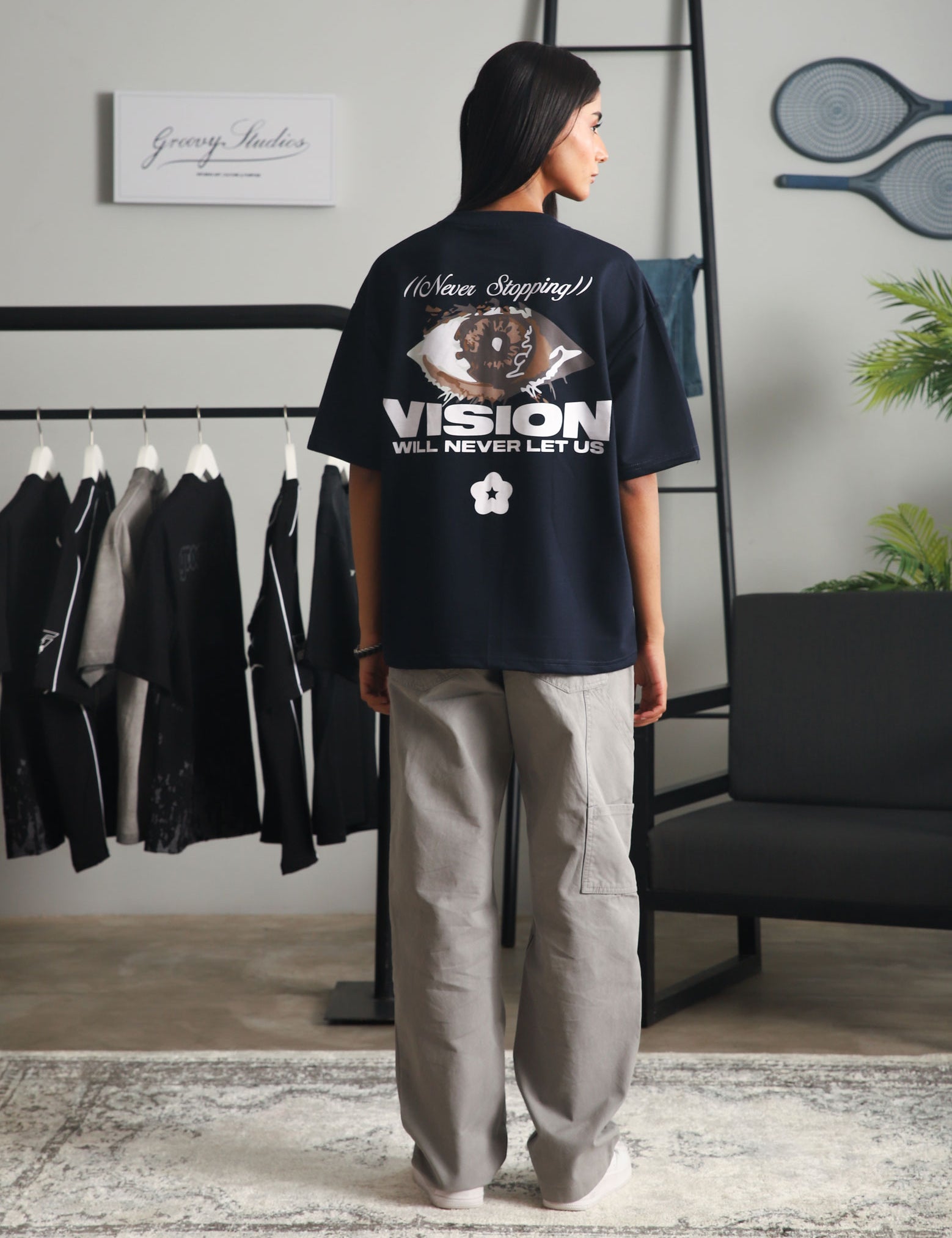 Back view of a woman wearing the "Never Stopping Culture" navy blue graphic t-shirt featuring an eye design and bold text, styled with light grey cargo pants in a minimalist studio setup.

