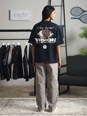 Back view of a woman wearing the "Never Stopping Culture" navy blue graphic t-shirt featuring an eye design and bold text, styled with light grey cargo pants in a minimalist studio setup.
