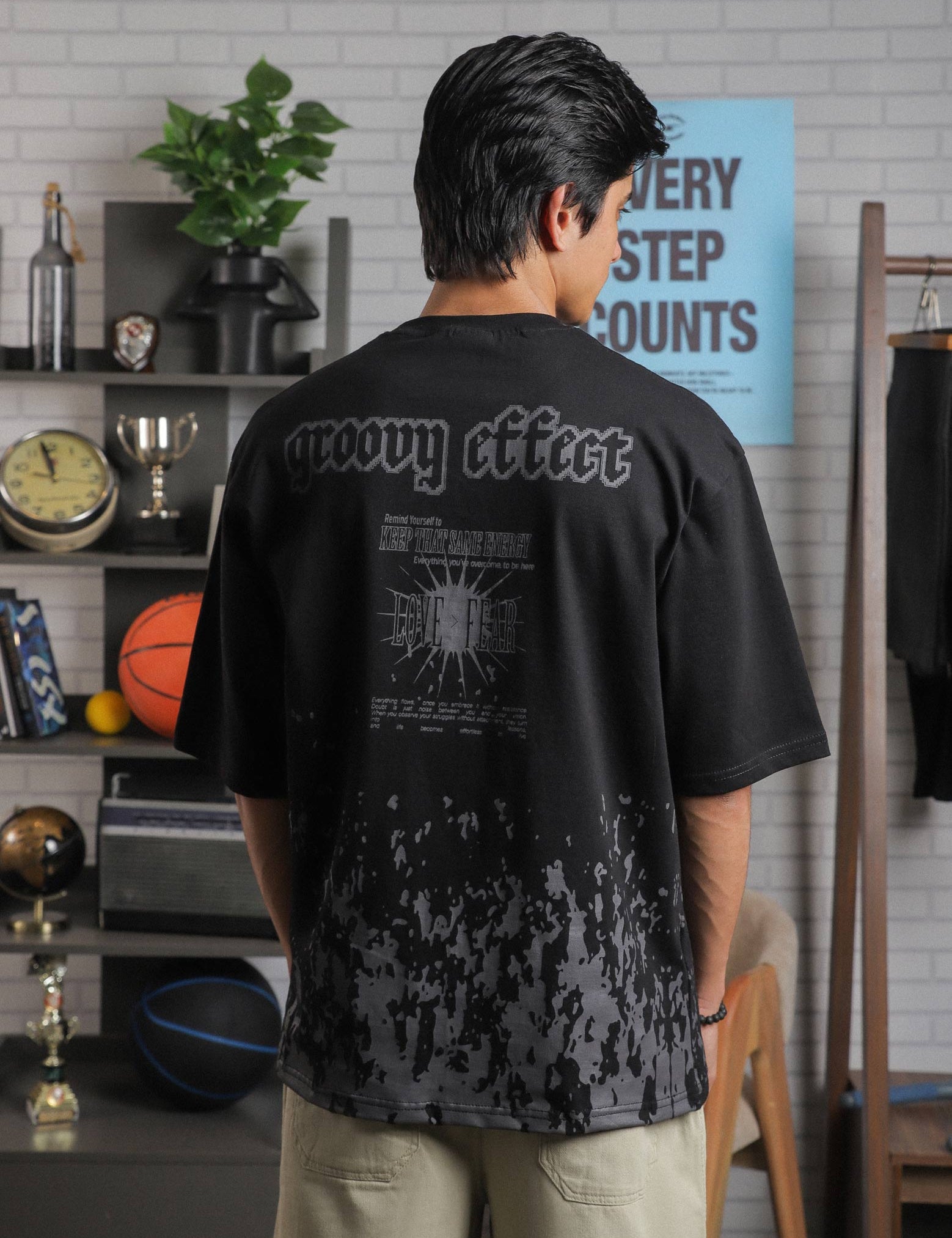 Back view of male model wearing “Groovy Effect” t-shirt in black, with bold screenprinted quote and “Love Over Fear” graphic in grey tones.