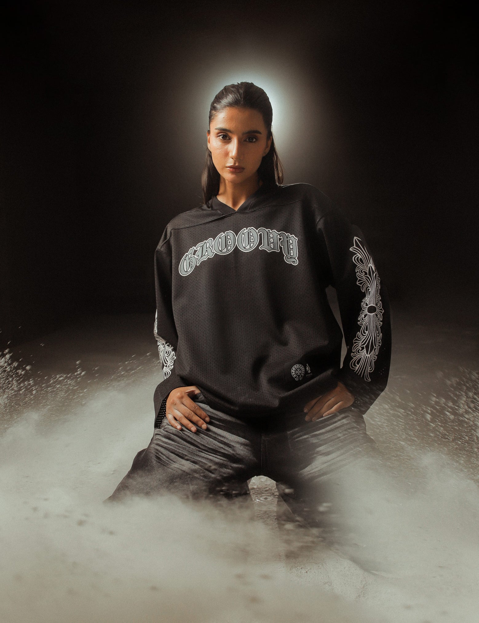 Female model in a dark, moody set wearing GROOVY’s Chrome Hearts-inspired black full sleeves jersey with gothic prints, surrounded by dramatic dust clouds for an ethereal streetwear campaign look.
