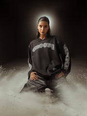 Female model in a dark, moody set wearing GROOVY’s Chrome Hearts-inspired black full sleeves jersey with gothic prints, surrounded by dramatic dust clouds for an ethereal streetwear campaign look.