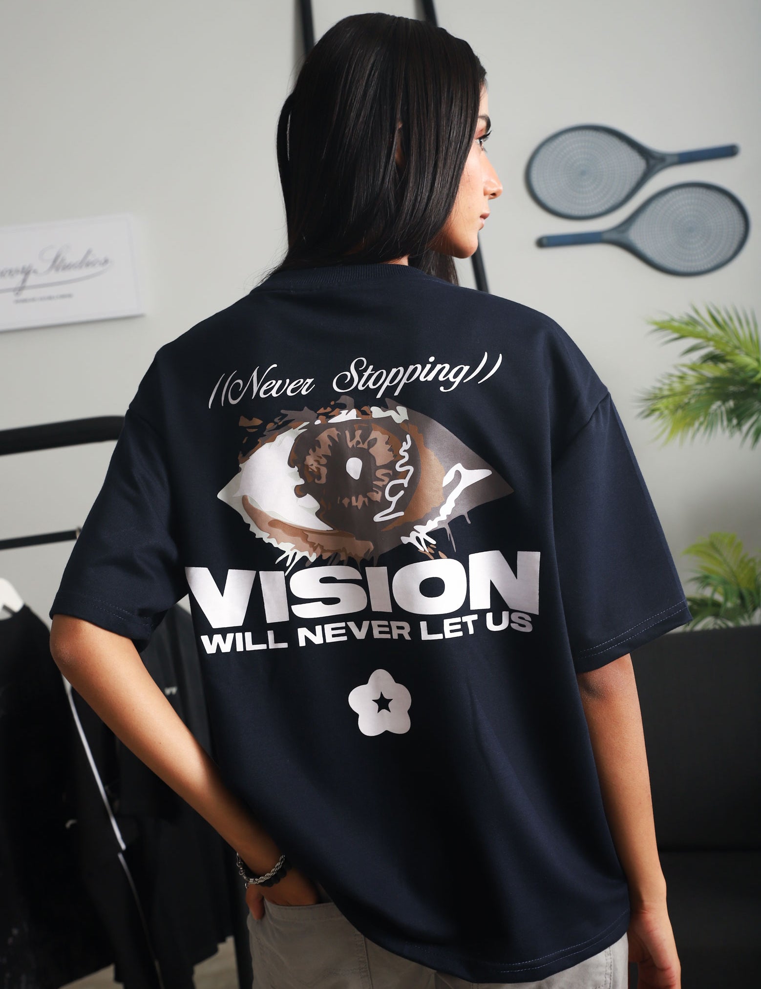 Close-up back view of a woman wearing the navy blue "Never Stopping Culture" t-shirt featuring a bold eye graphic and motivational slogan, styled with light grey pants in a contemporary studio space.

