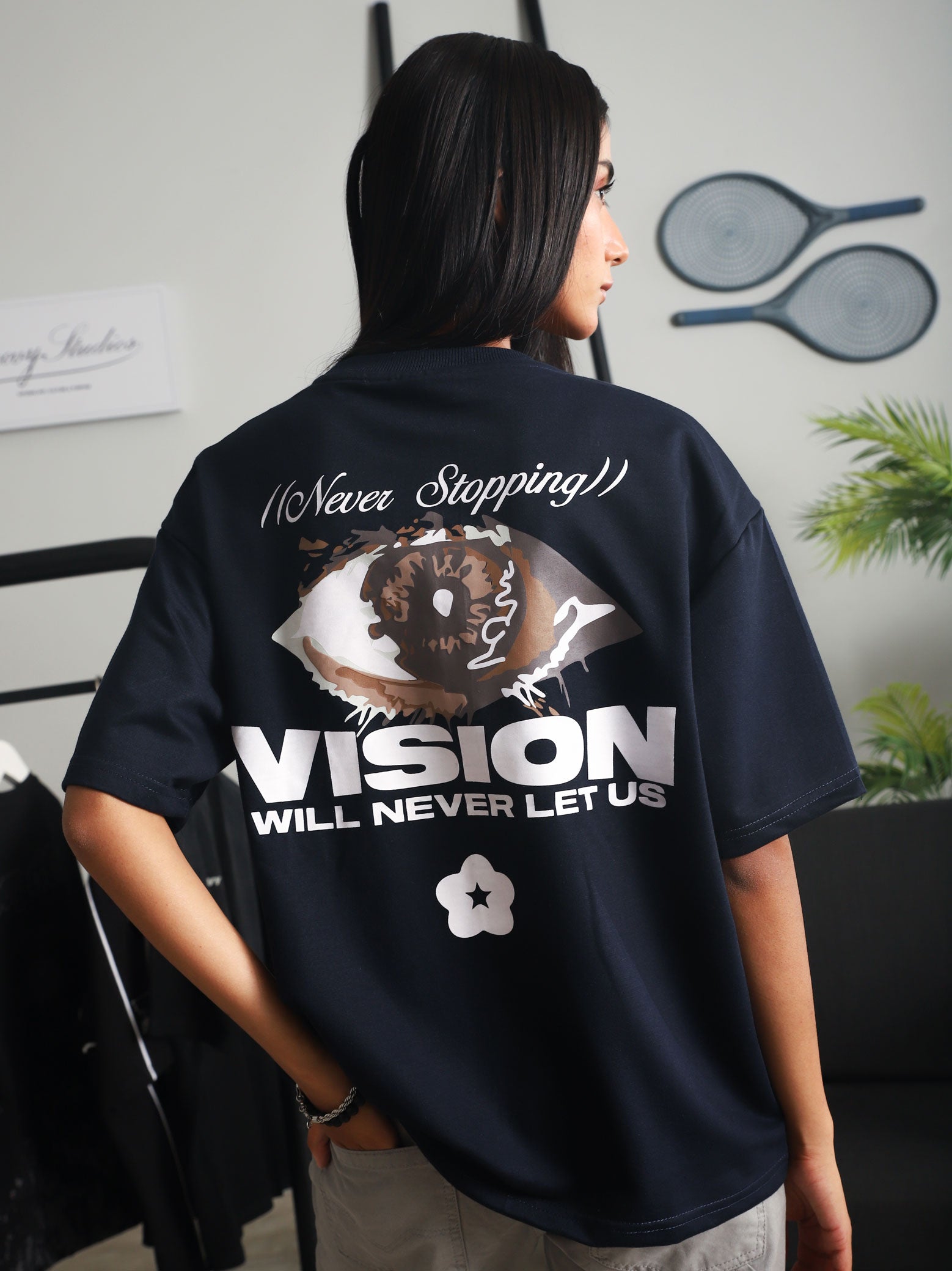 Close-up back view of a woman wearing the navy blue "Never Stopping Culture" t-shirt featuring a bold eye graphic and motivational slogan, styled with light grey pants in a contemporary studio space.
