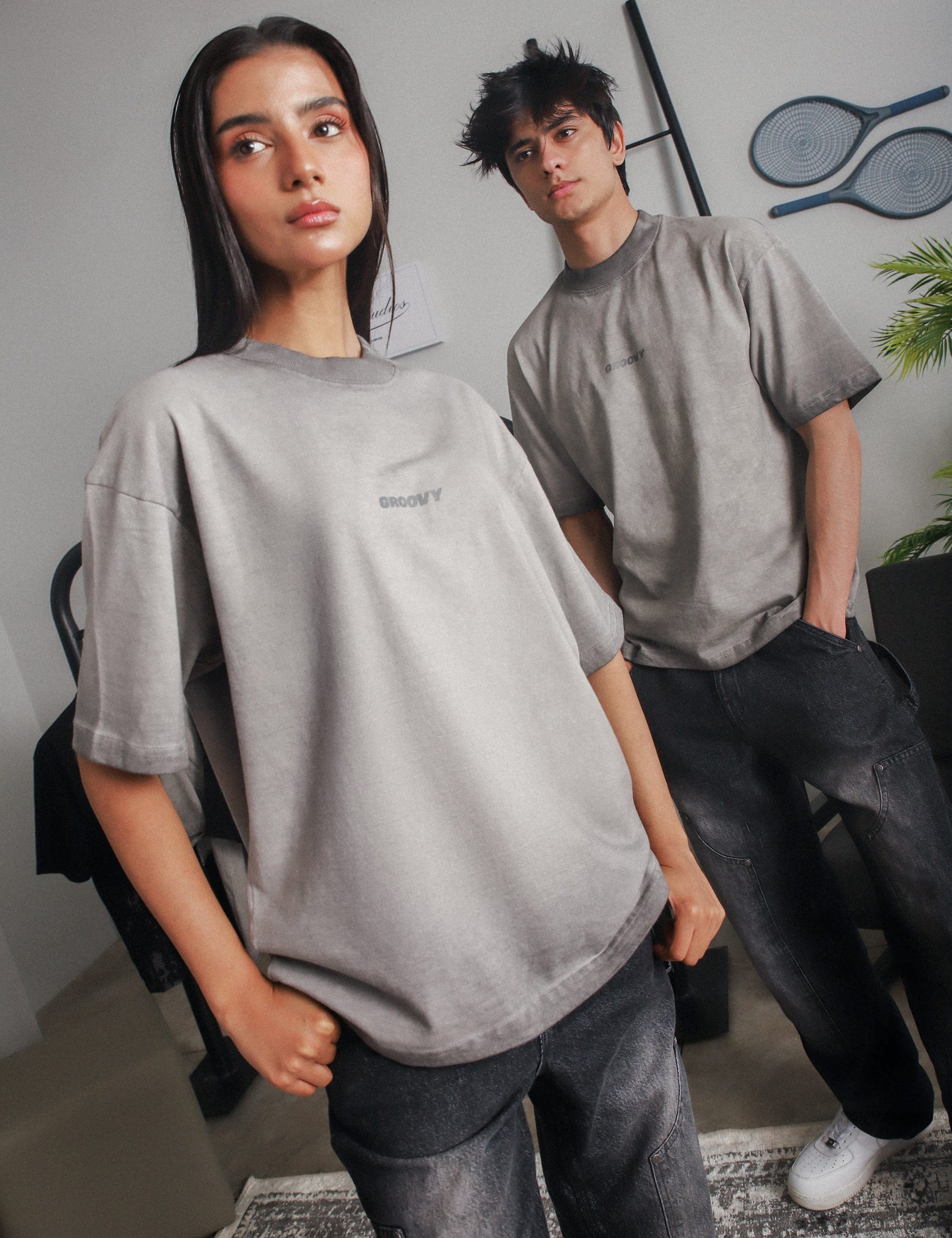 Man and woman wearing GROOVY Oil Fade Blank t-shirts styled with faded black jeans, shot in a minimal indoor setting with streetwear pieces in the background.

