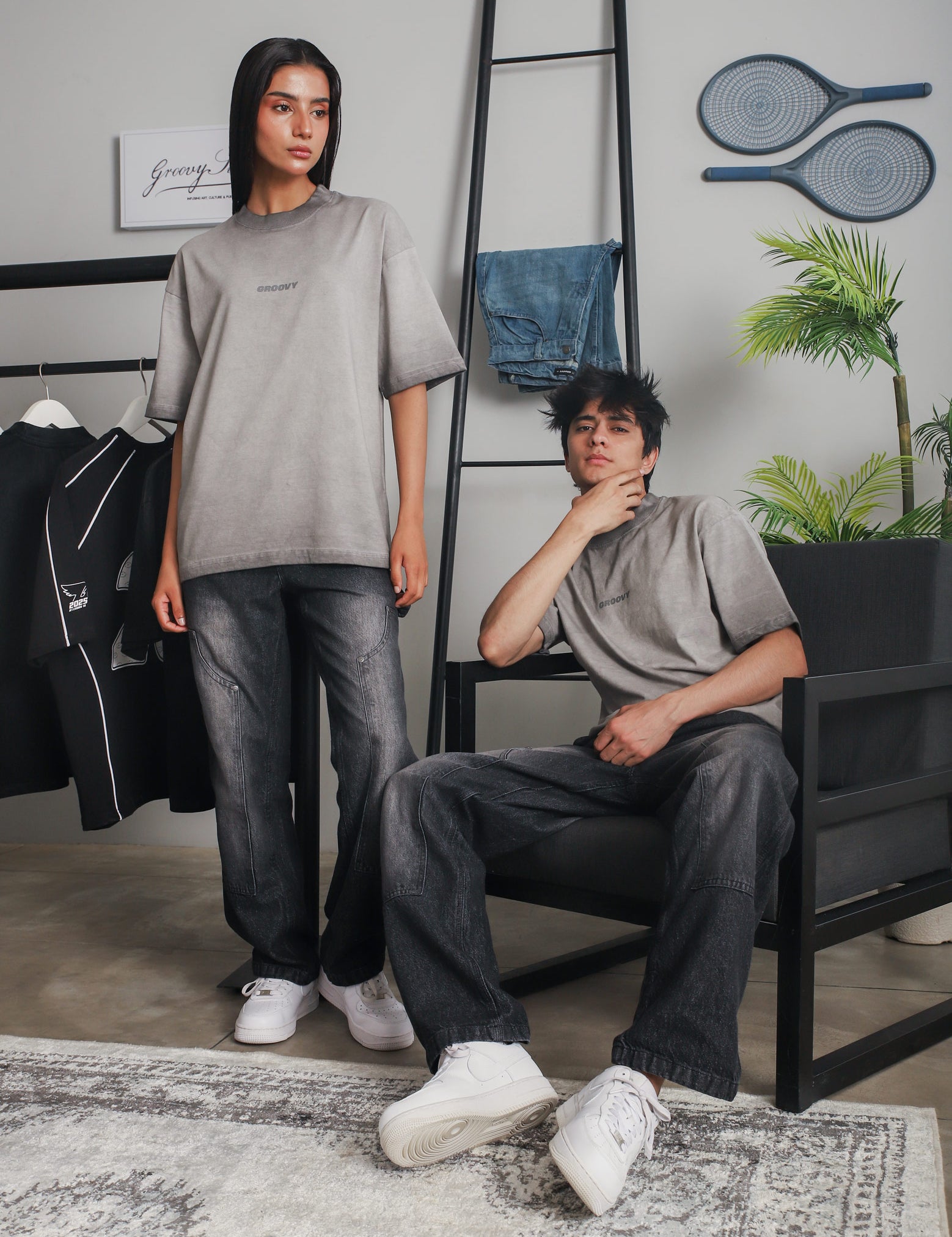 Man and woman wearing GROOVY Oil Fade Blank t-shirts styled with faded black jeans, shot in a minimal indoor setting with streetwear pieces in the background.
