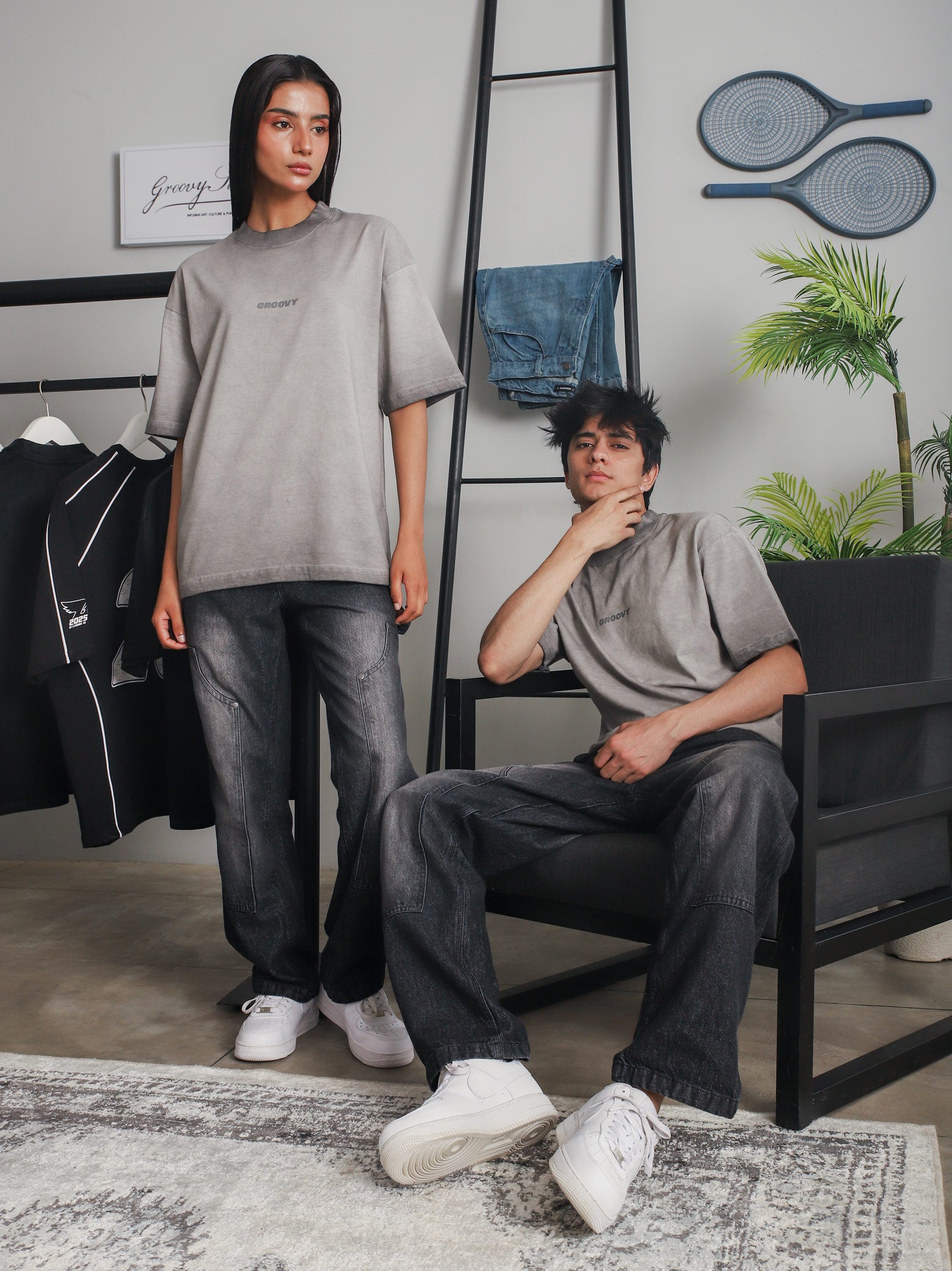Man and woman wearing GROOVY Oil Fade Blank t-shirts styled with faded black jeans, shot in a minimal indoor setting with streetwear pieces in the background.
