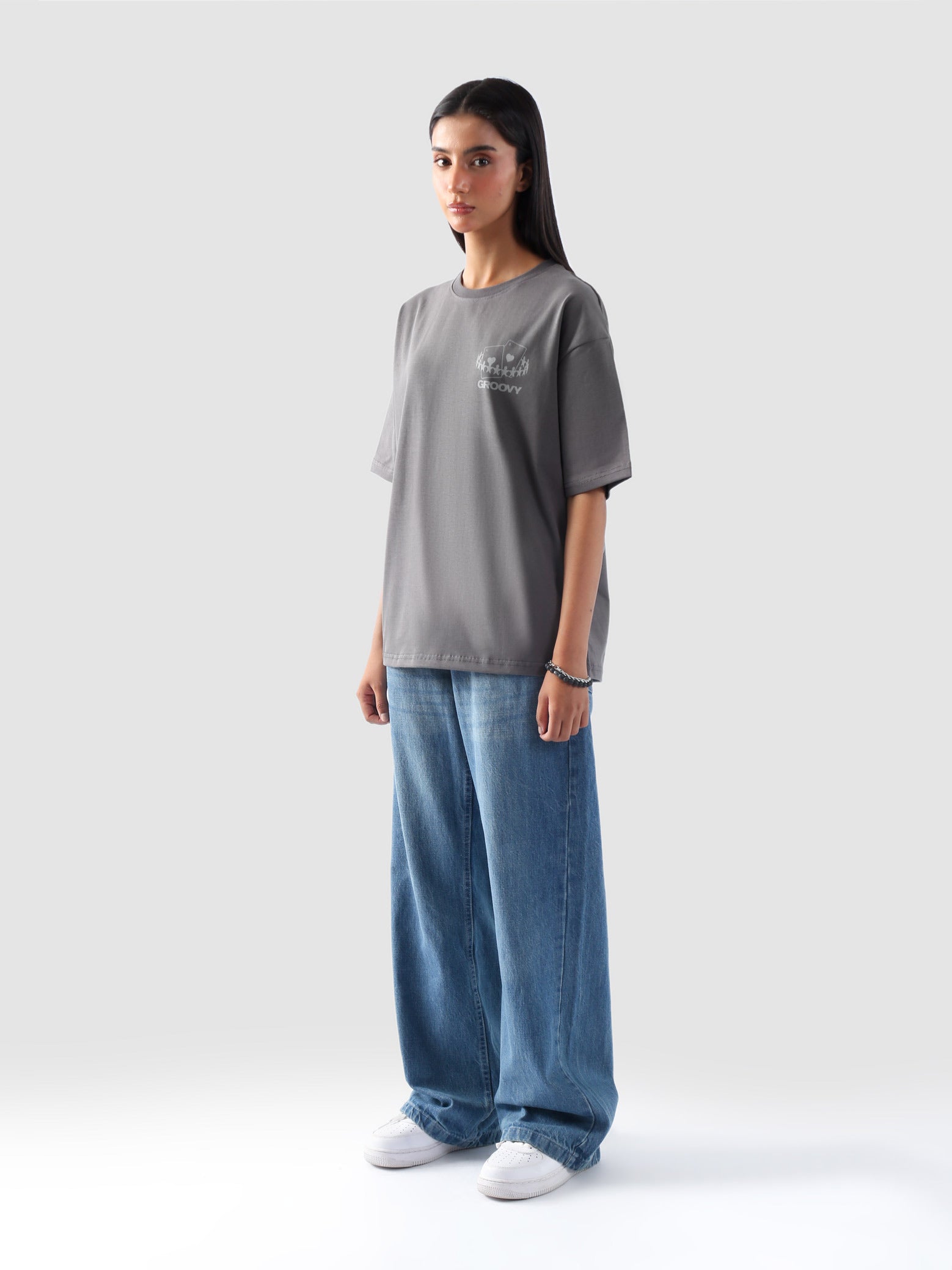 Model wearing stone grey oversized t-shirt for women in Pakistan, styled with wide-leg blue denim jeans – Groovy streetwear graphic tee with relaxed fit and drop shoulder design