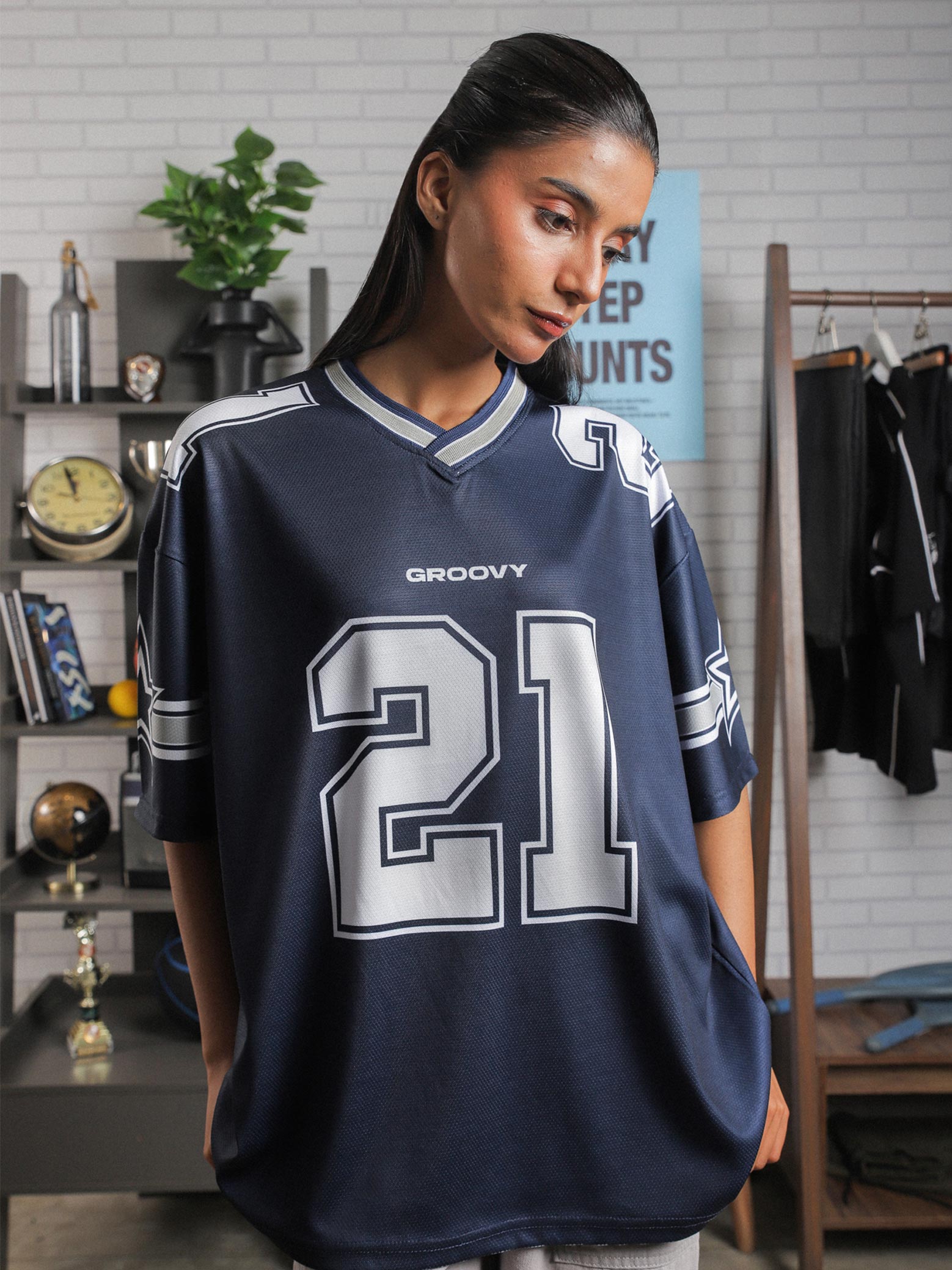 Female model in relaxed pose wearing GROOVY jersey, positioned near motivational poster and sports shelf setup.
