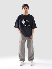 Front-facing shot of a male model wearing the "Never Stopping Culture" navy blue tee with bold white CLTR star graphic, paired with light grey baggy cargo pants and white sneakers, set against a minimalist grey backdrop.