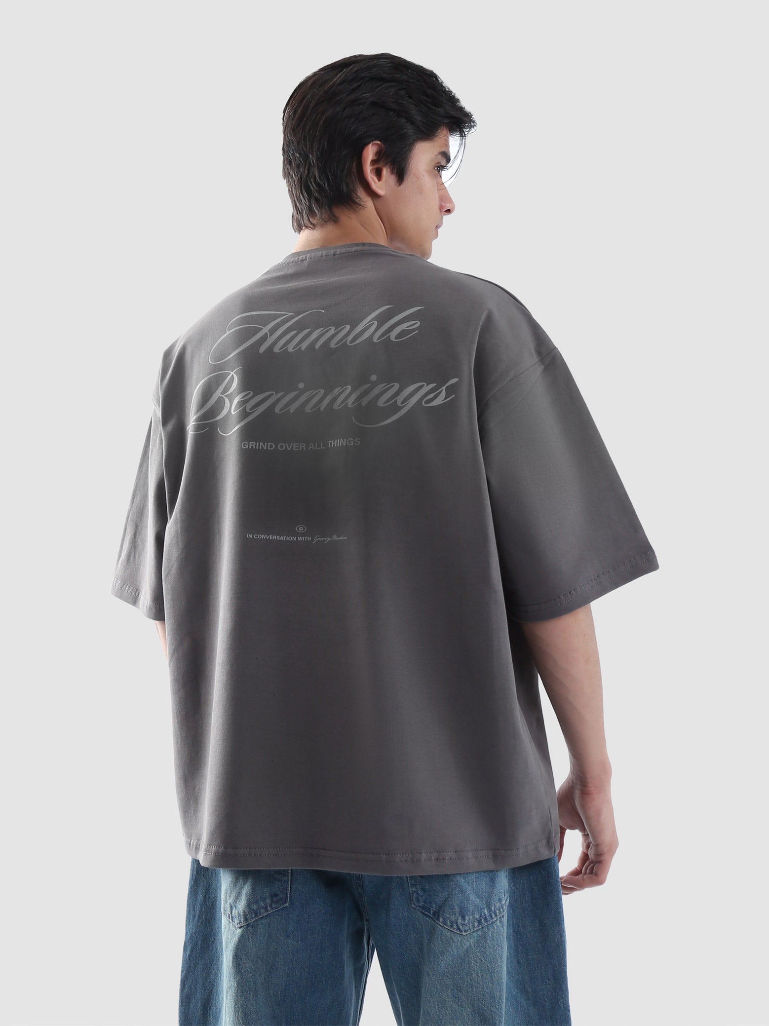 Back view of male model wearing stone grey oversized t-shirt with 'Humble Beginnings' graphic text – streetwear tee in Pakistan by Groovy, styled with loose denim jeans