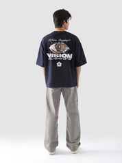 Back view of a male model wearing the "Never Stopping Culture" navy blue t-shirt, showcasing the large puff and screen-printed eye graphic with the slogan “VISION WILL NEVER LET US,” symbolizing determination and relentless ambition.