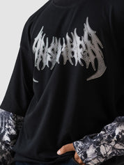 A close-up of a person wearing a black oversized graphic t-shirt with a metallic silver 'The Ashes' logo across the chest, layered over a black and white patterned long-sleeve shirt.